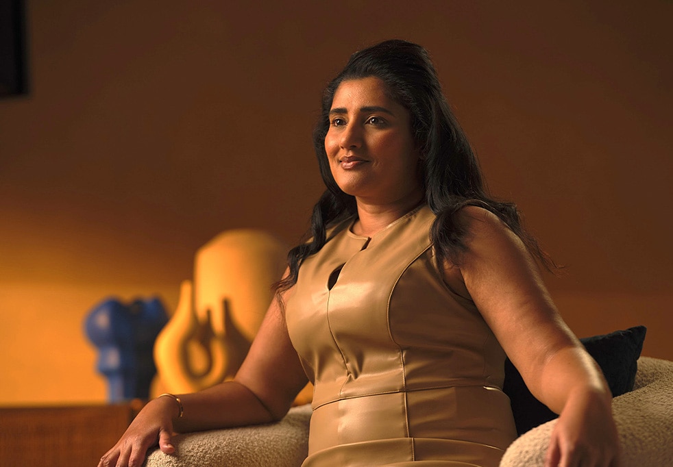 Tanusta's photo with long dark hair, wearing a tan dress, sitting on a chair and smiling slightly