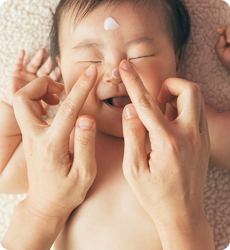 a naked baby smiling with two fingers pointed at the baby with a drop of lotion on the baby's forehead